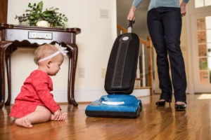 A mother vacuums with a Riccar Cordless SupraLite while her child watches. Contact or call Stark's Vacuums if you need a central vacuum replacement or upgrade.